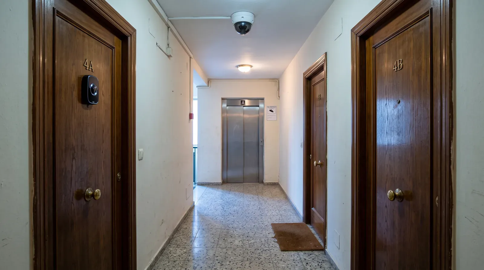 Rellano de un edificio de viviendas español con suelo de terrazo y puertas de madera enfrentadas a pocos metros: sobre la puerta 4A, una mirilla digital negra con lente visible orientada al pasillo; en el techo del descansillo, una cámara domo de videovigilancia de la comunidad, y junto al ascensor el cartel informativo de zona videovigilada.